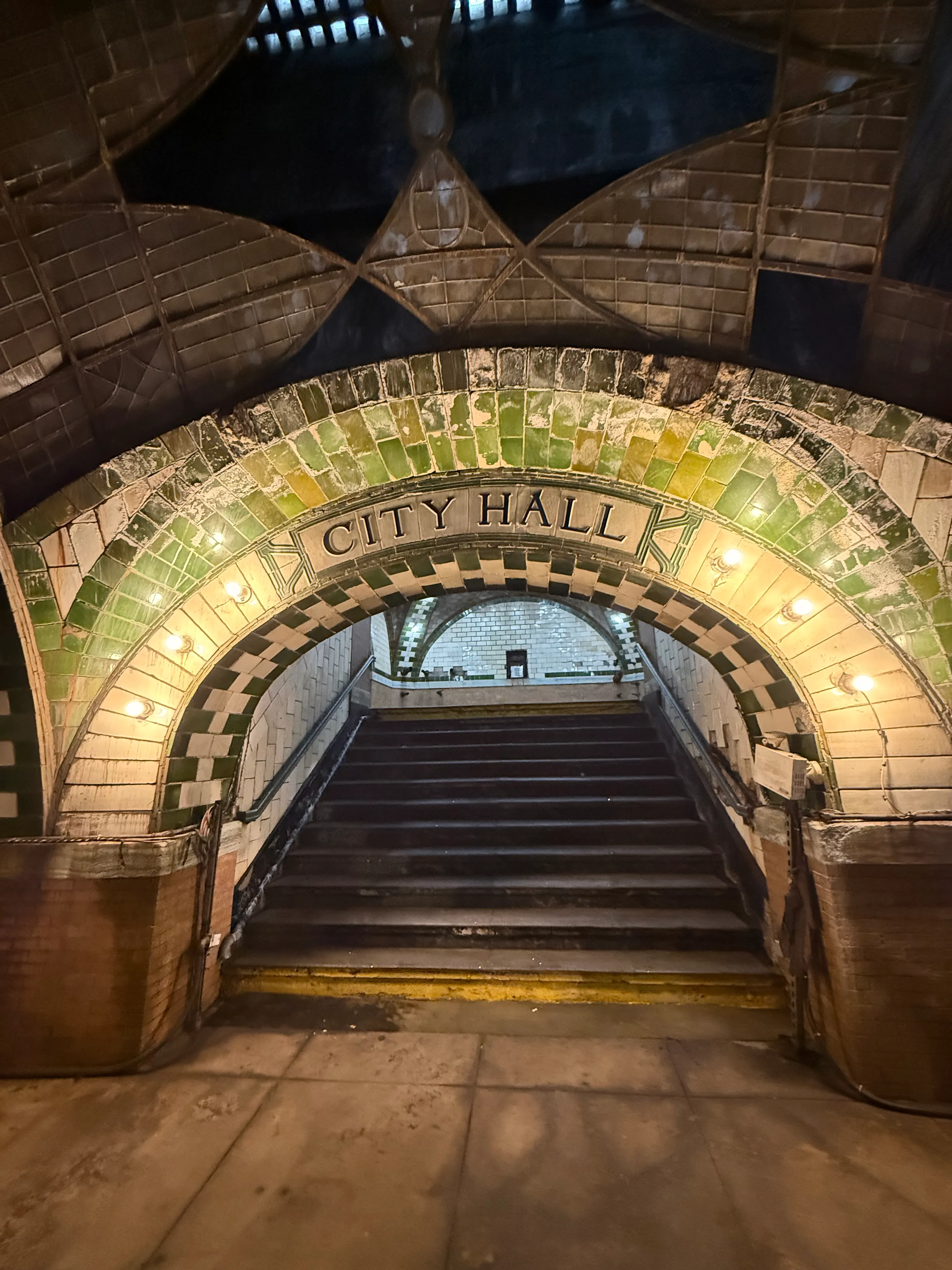 Beautiful, old station sign saying "City Hall" in front of the stairs going up and out of the station. The words are placed in a green-and-white tile arch with embedded lightbulbs. There's a hint the main platform skylight visible as well.